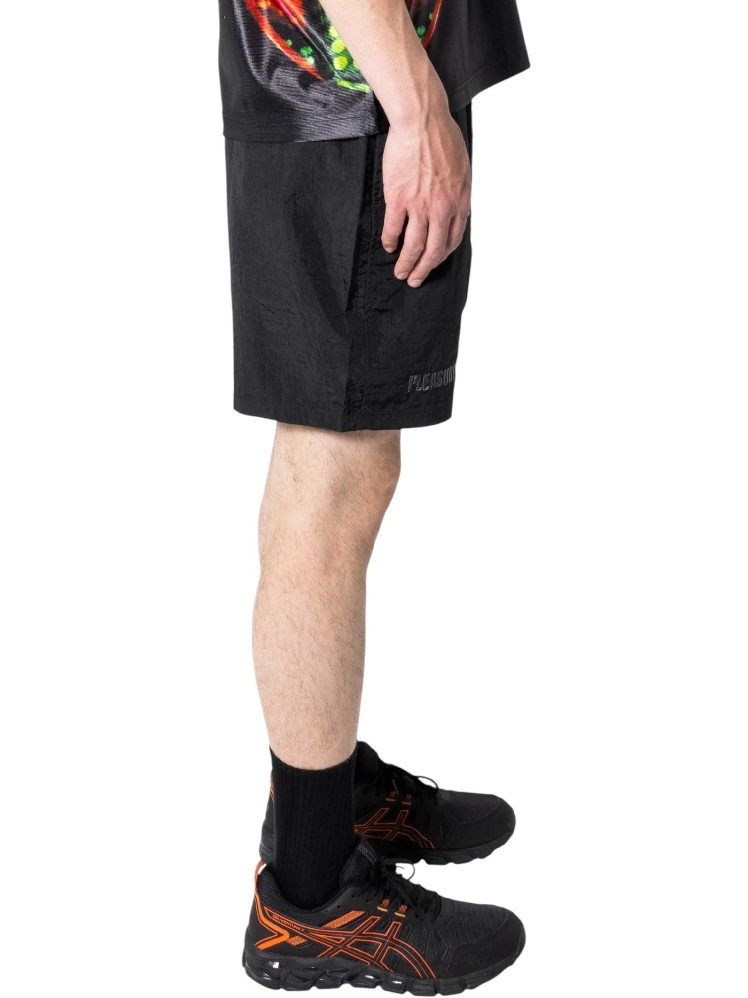 A person stands in profile wearing PLEASURES Crush Hiking Shorts Black with an integrated belt, a black shirt with a colorful design, black socks, and black sneakers featuring orange accents.