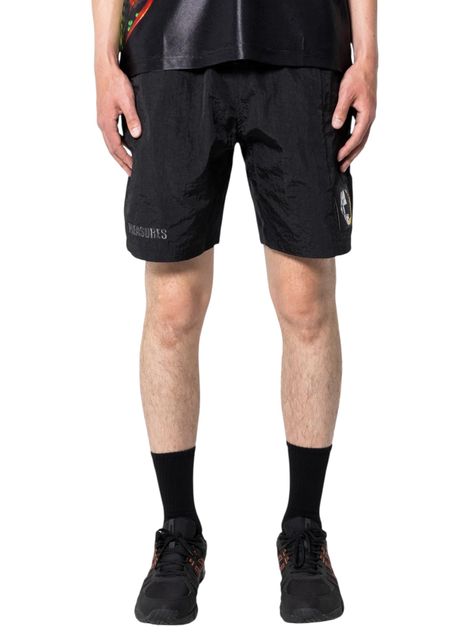 Someone stands against a plain background wearing PLEASURES Crush Hiking Shorts Black—nylon shorts with an elasticized waistband and small graphic on one leg—paired with black socks and sneakers.