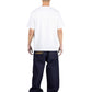 A person with a shaved head stands facing away, wearing the PLEASURES Cut Here Heavyweight T-Shirt in white and loose-fitting dark blue jeans featuring yellow stitching on the back pockets.
