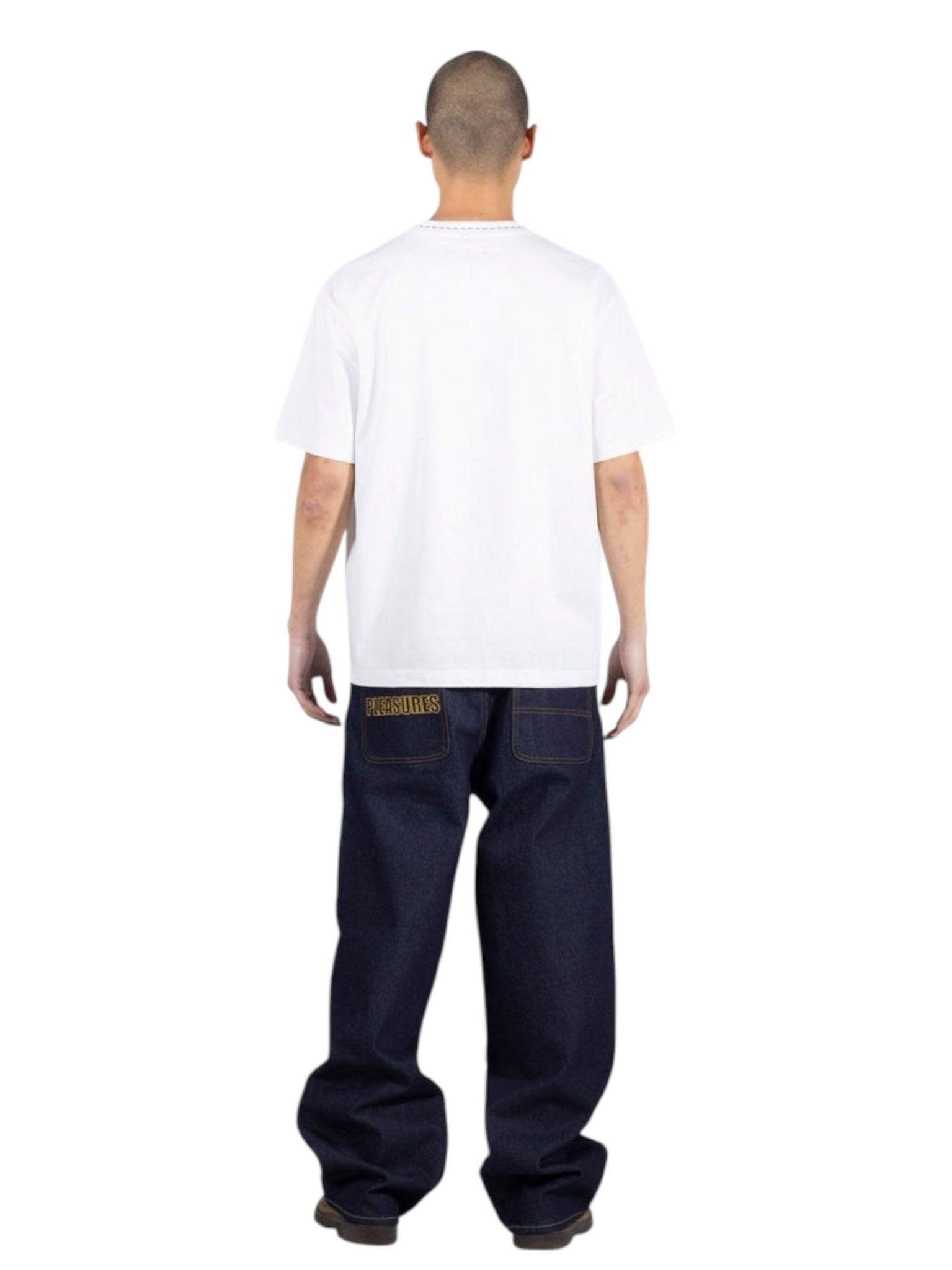 A person with a shaved head stands facing away, wearing the PLEASURES Cut Here Heavyweight T-Shirt in white and loose-fitting dark blue jeans featuring yellow stitching on the back pockets.