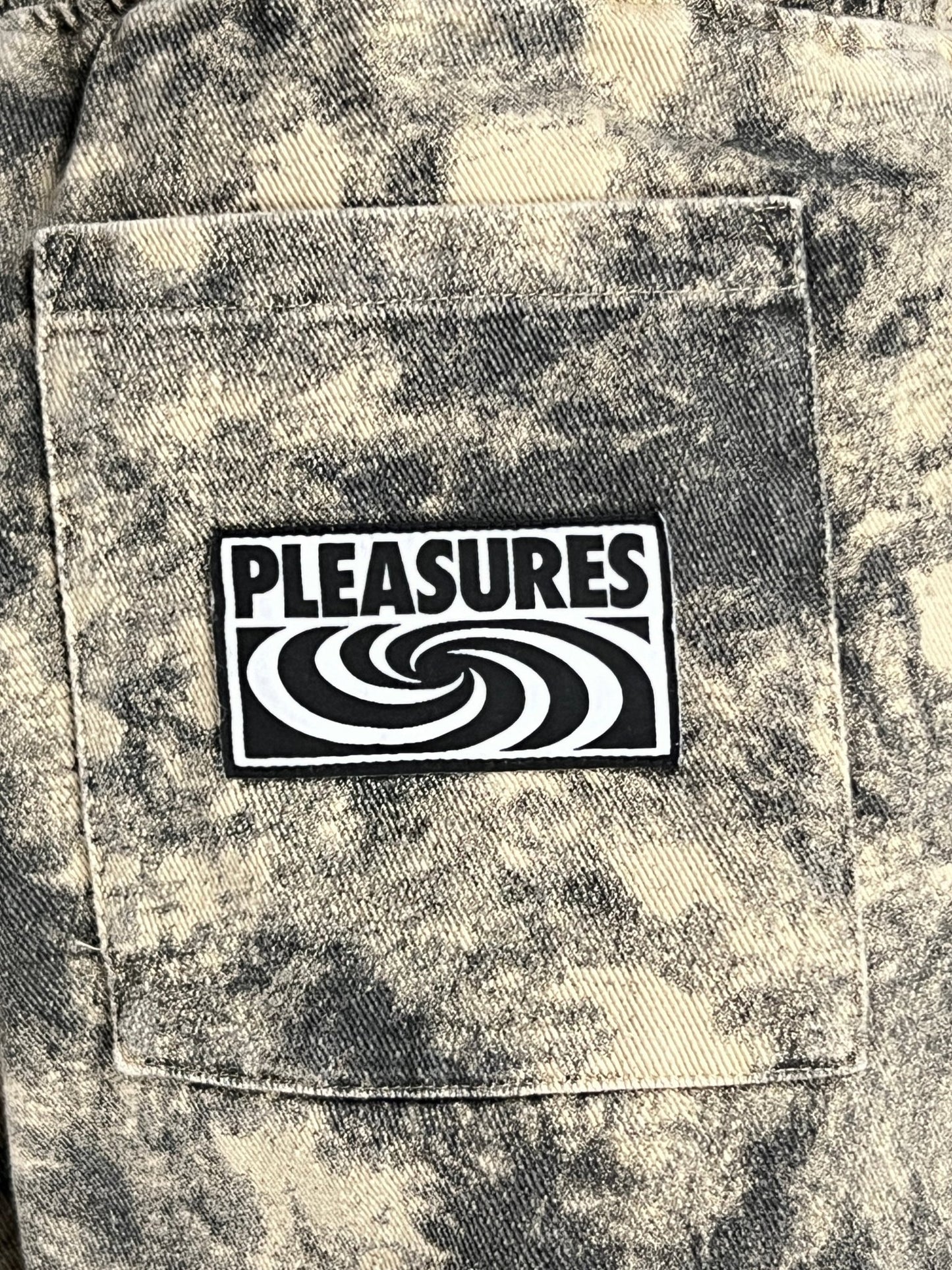 Close-up of a denim pocket with a black and white patch reading "PLEASURES" on it. The 100% cotton denim has a marbled, acid-washed appearance reminiscent of the PLEASURES CYCLONE SHORTS BLACK collection.