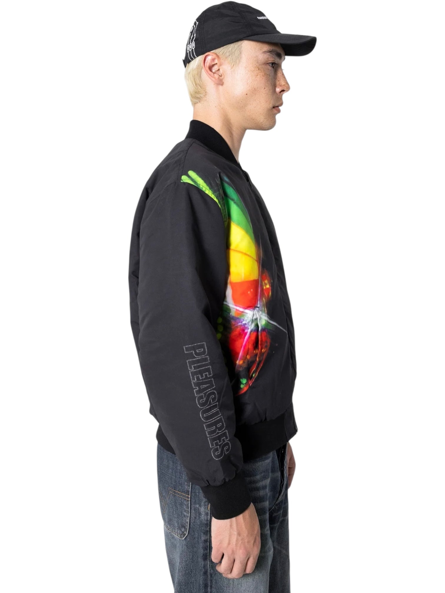 A person in a black cap, Pleasures Daft Club Reversible Bomber Black by PLEASURES with sublimated graphics and sleeve lettering, and blue jeans stands facing sideways.
