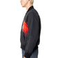 A person in profile wears the PLEASURES Daft Club Reversible Bomber Black with a red graphic on the front, paired with a black cap and blue jeans, standing against a white background.