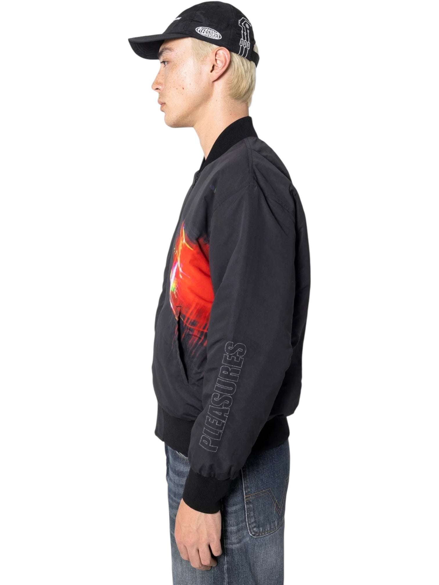 A person in profile wears the PLEASURES Daft Club Reversible Bomber Black with a red graphic on the front, paired with a black cap and blue jeans, standing against a white background.