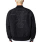 A person wearing the PLEASURES Daft Club Reversible Bomber Black jacket with a list of words on the back and a black cap, shown from behind.