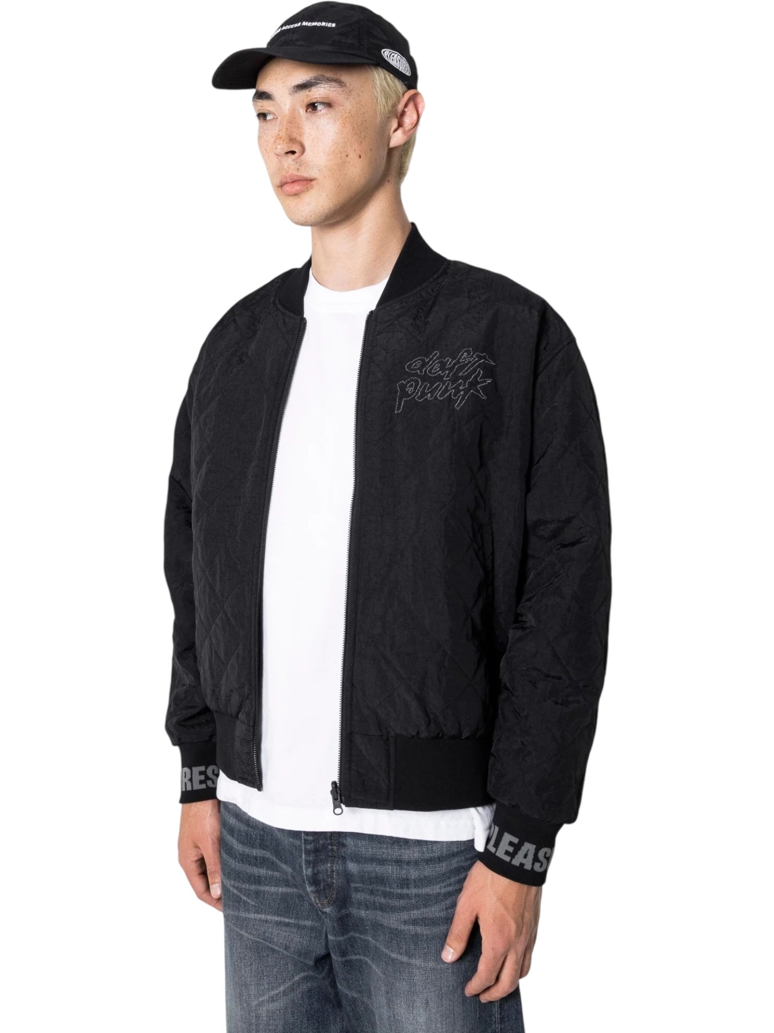 A person wearing the PLEASURES Daft Club Reversible Bomber Black, a white t-shirt, jeans, and a black cap stands against a white background.