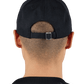 A person with short hair wears a backwards Pleasures Daniele Cap Black, showcasing its adjustable fit and an embroidered boar logo that marks the exclusive PLEASURES x Iuter collaboration.