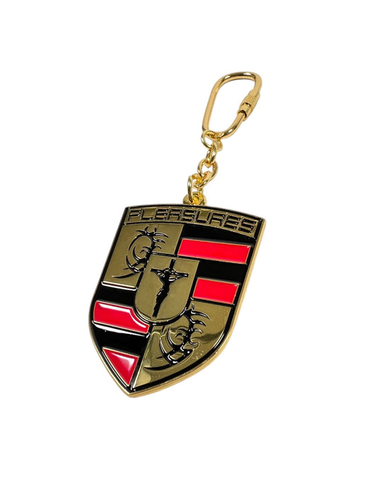 Gold and black PLEASURES Fast Life Keychain Multi with a shield design features a horse, red and black stripes, and text celebrating life's small elegant pleasures.