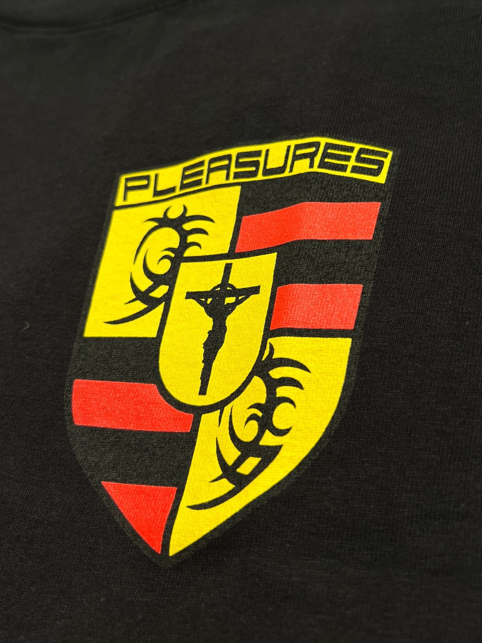The Pleasures Fast Life T-shirt Black by PLEASURES features a boxy fit, black cotton fabric, and a yellow and red crest with “PLEASURES,” tribal patterns, and a crucifixion silhouette at the center.