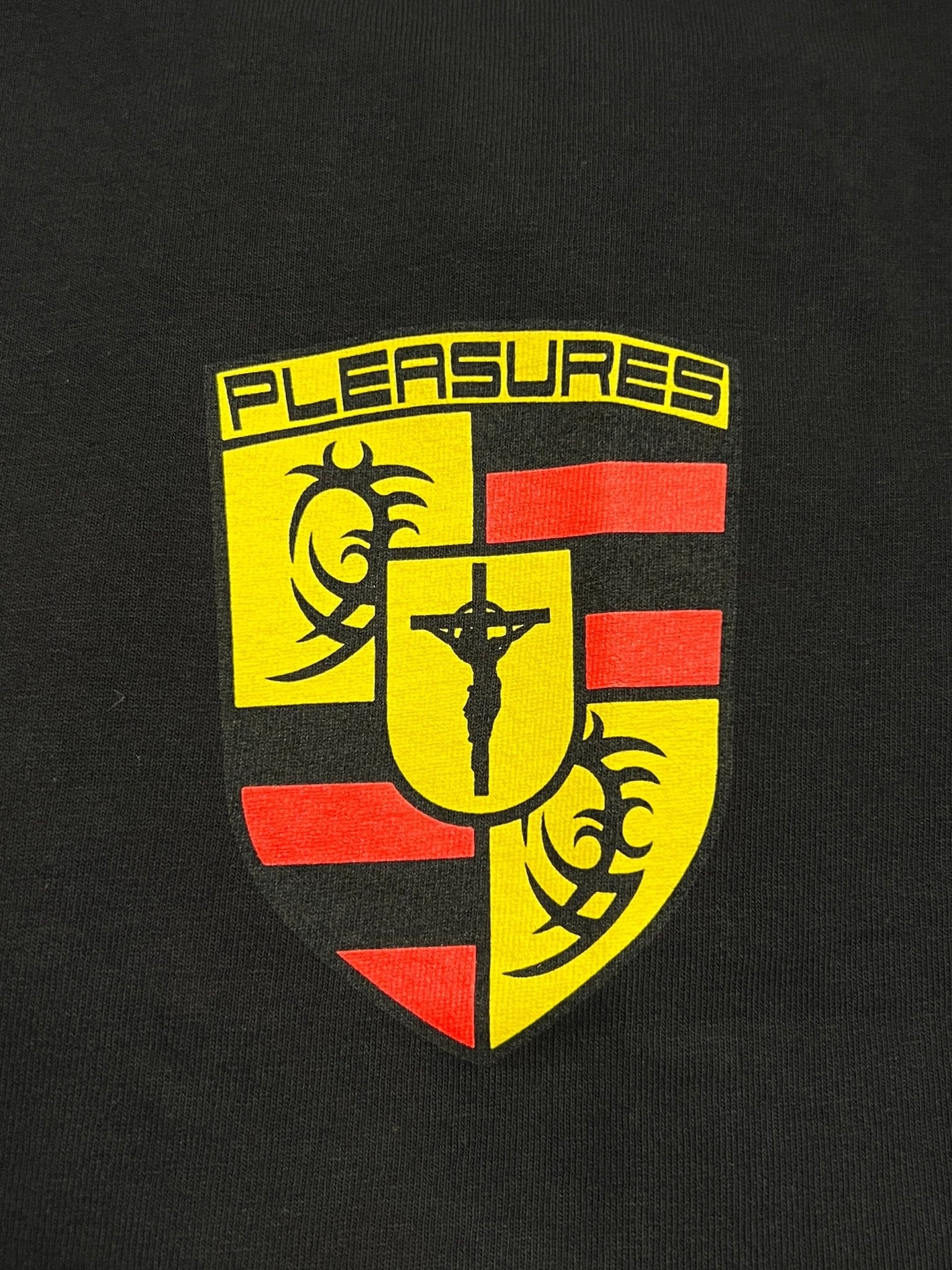 The Pleasures Fast Life T-shirt Black by PLEASURES features a yellow and red shield logo with "PLEASURES" at the top, tribal designs, and a crucifix, printed on garment-dyed black cotton in a boxy fit.