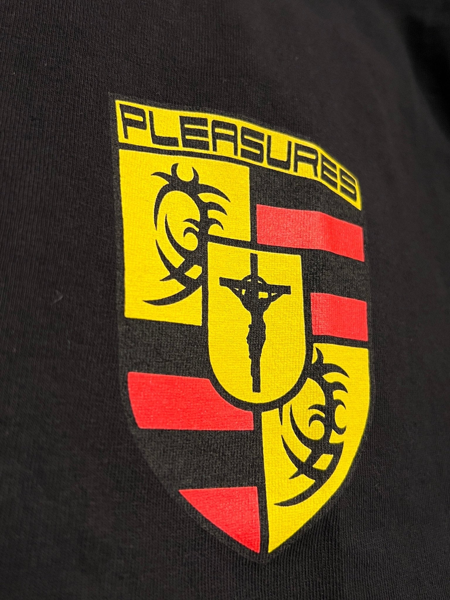The Pleasures Fast Life T-shirt Black by PLEASURES is a black, boxy fit shirt with a yellow and red crest, “PLEASURES” at the top, and a central crucifixion graphic.