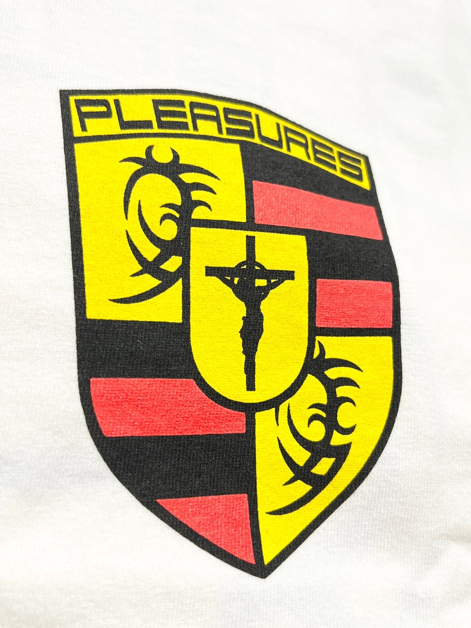 The Pleasures Fast Life T-shirt White by PLEASURES features a crest-style emblem with red/yellow blocks, black tribal designs, "PLEASURES" at the top, and a crucifixion figure on garment dyed cotton. Made in the USA.