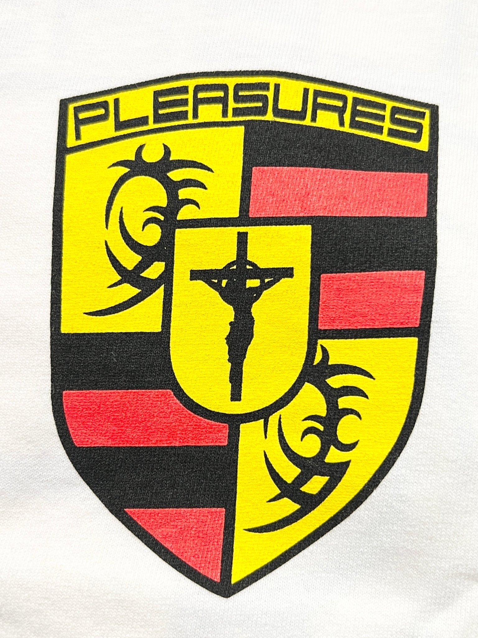 The Pleasures Fast Life T-shirt White by PLEASURES features a yellow and red shield logo with black tribal designs, a central crucifix, and "PLEASURES" at the top, printed on soft cotton and made in the USA.