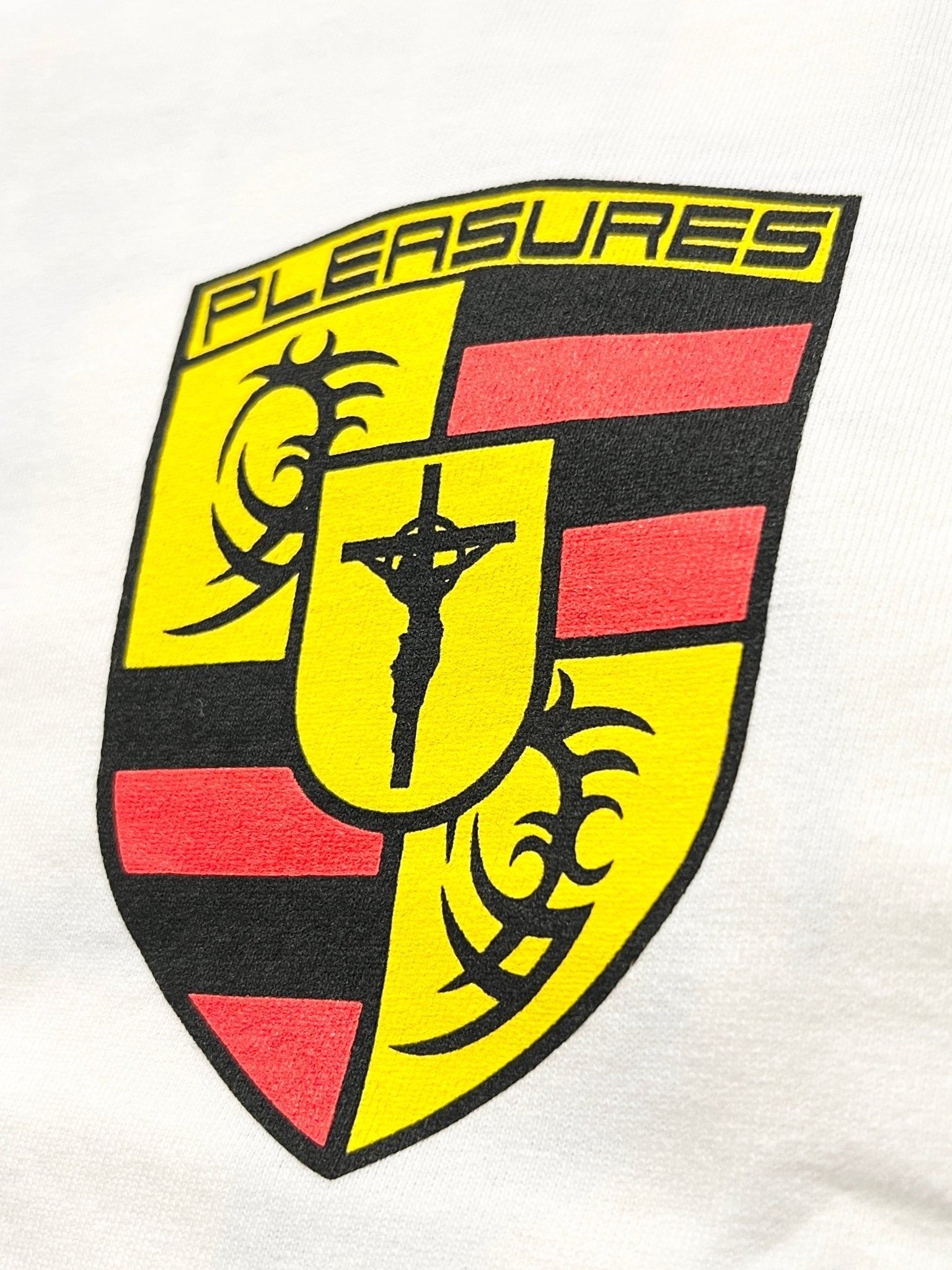 The Pleasures Fast Life T-shirt in white features a yellow and red shield logo with "PLEASURES," tribal designs, and a crucifix silhouette on garment-dyed cotton for a unique finish.