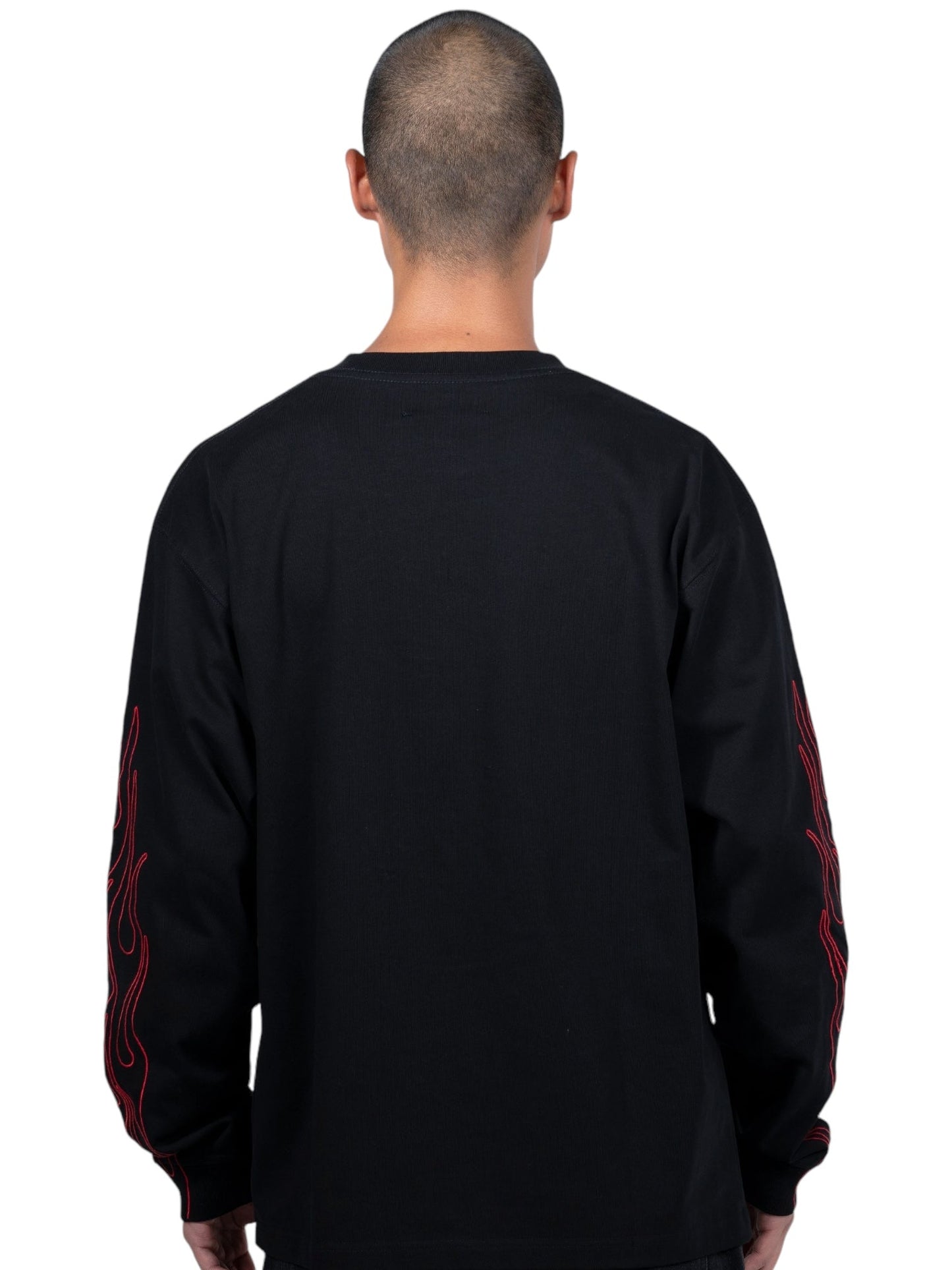 A person with a shaved head, facing away, is wearing the PLEASURES Fiery Heavyweight L/S T-shirt in Black, featuring red flame designs on the sleeves and an embroidered logo.