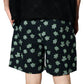 A person is seen from behind wearing PLEASURES Floral Seersucker Shorts Black, featuring a green and gray floral pattern. Their hands are at their sides, with part of a black top also visible.