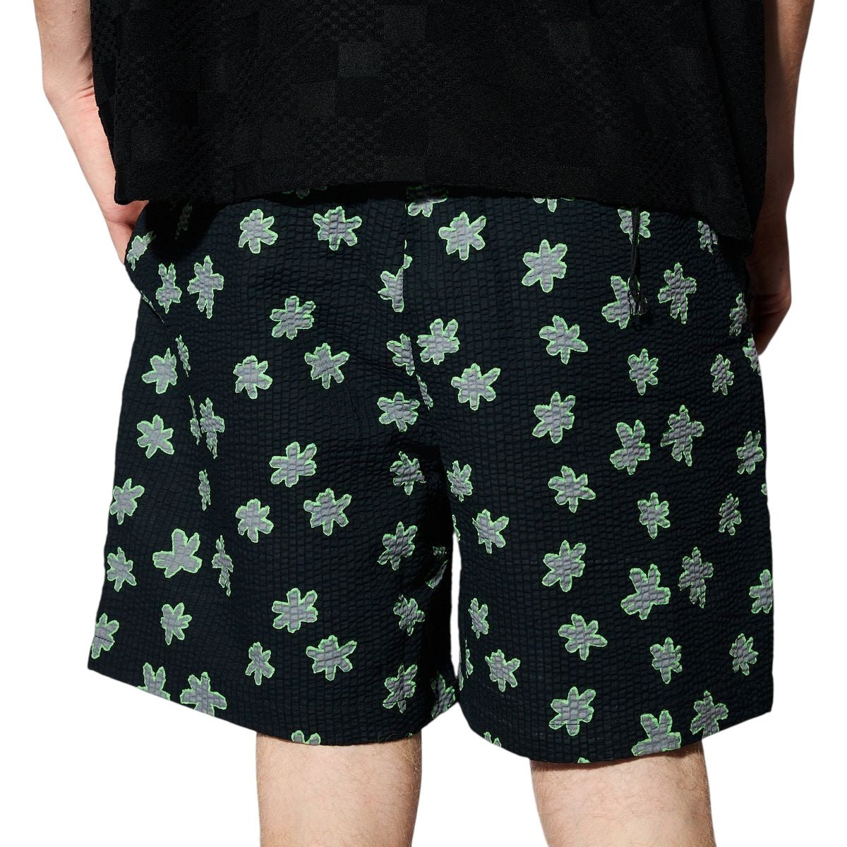 A person is seen from behind wearing PLEASURES Floral Seersucker Shorts Black, featuring a green and gray floral pattern. Their hands are at their sides, with part of a black top also visible.