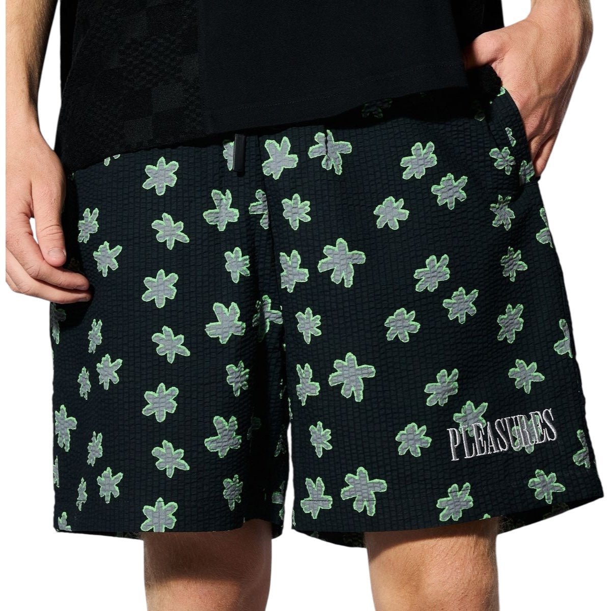 Someone wearing the PLEASURES Floral Seersucker Shorts Black, featuring a green floral pattern and "PLEASURES" embroidered in white on the left leg.