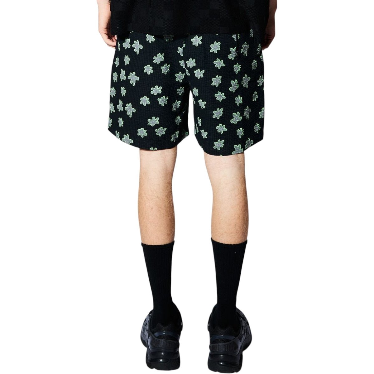Someone wearing PLEASURES Pleasures Floral Seersucker Shorts Black with a green four-leaf clover pattern, paired with black socks and shoes, photographed from behind against a white background.