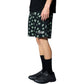 A person wearing PLEASURES Floral Seersucker Shorts Black, black socks, black sneakers, and a black shirt with an embroidered logo stands against a white background.