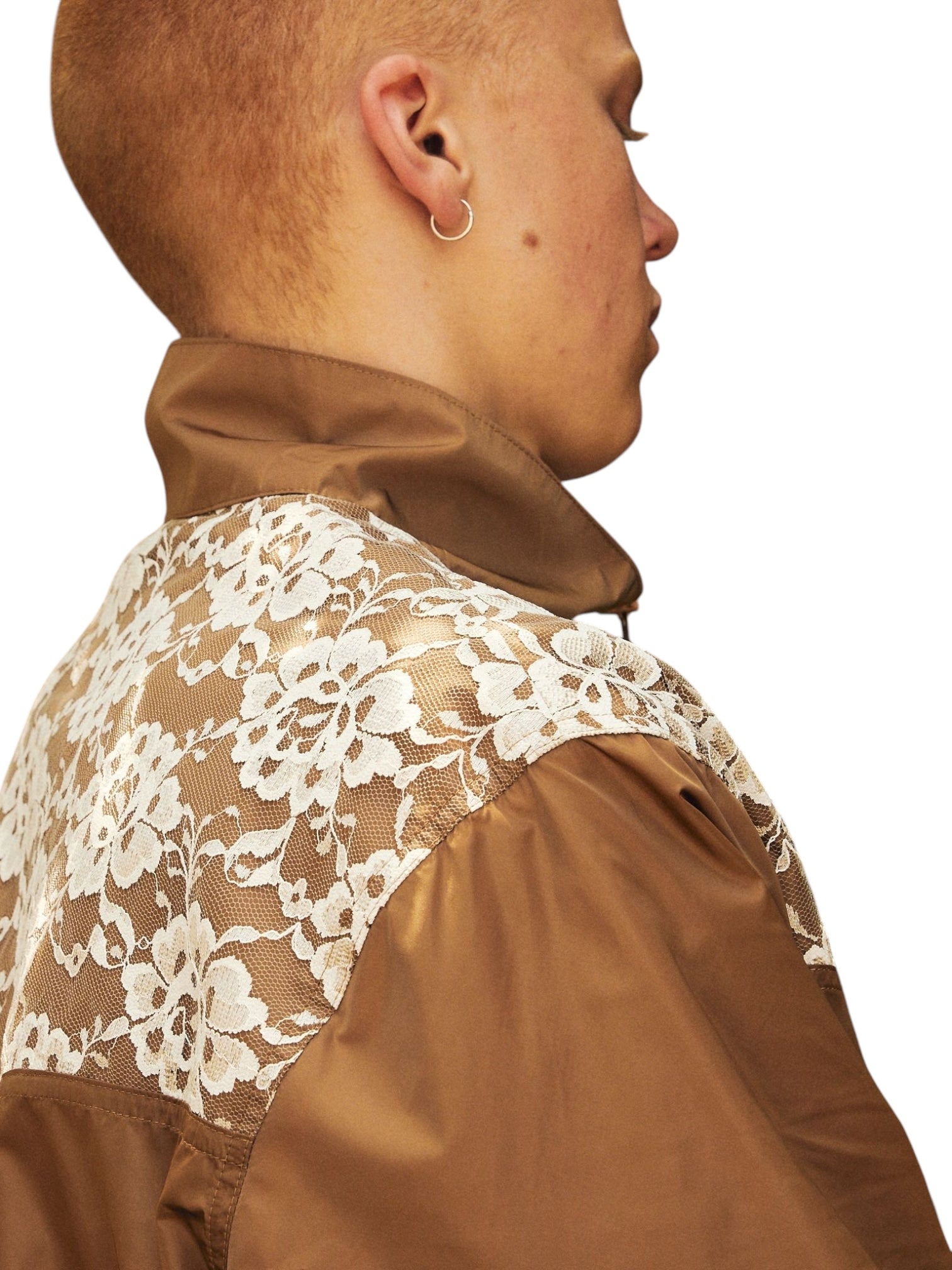 A person with short hair wears the Pleasures Francesca Track Jkt Brown by PLEASURES, a chic brown jacket made from Polyamide, featuring an elegant white floral lace design on the shoulder and back, embodying Italian craftsmanship.
