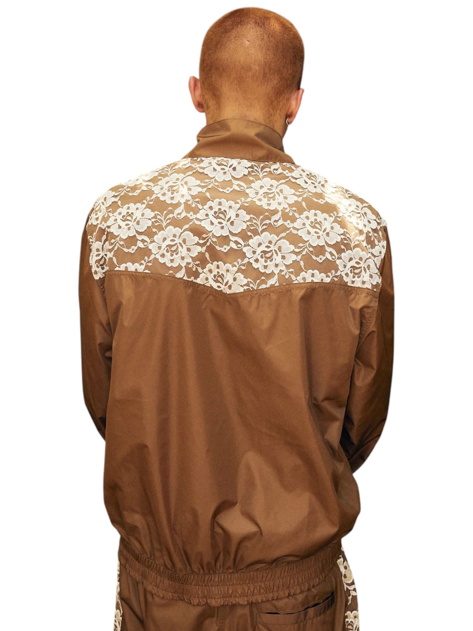 The person is wearing a chic Pleasures Francesca Track Jkt Brown by PLEASURES, featuring elegant white lace detailing on the back, against a plain background.