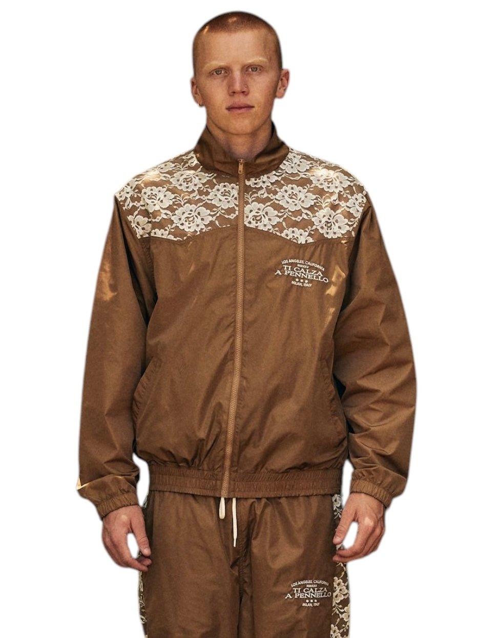 The individual is wearing the Pleasures Francesca Track Jkt Brown by PLEASURES, a brown Italian polyamide tracksuit with white lace accents and text, set against a plain background.