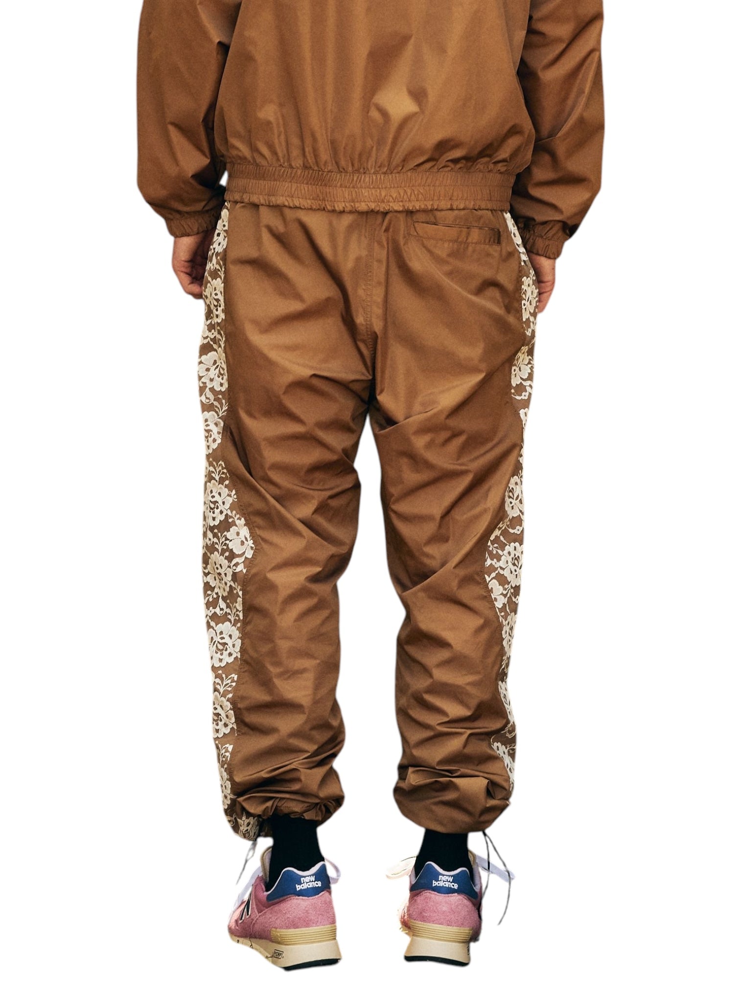 A fashionable person wears Pleasures Francesca Track Pants Brown by PLEASURES, featuring elegant lace side patterns, made from luxurious polyamide. Paired with pink and blue sneakers, the outfit is both stylish and functional against a white backdrop.