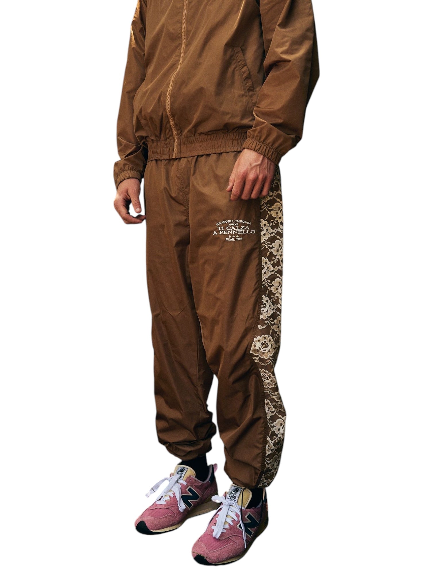 Wearing the Pleasures Francesca Track Pants in brown from PLEASURES, featuring patterned side stripes, this outfit is paired with pink sneakers. Made with durable polyamide, the track pants blend Italian elegance and functionality.