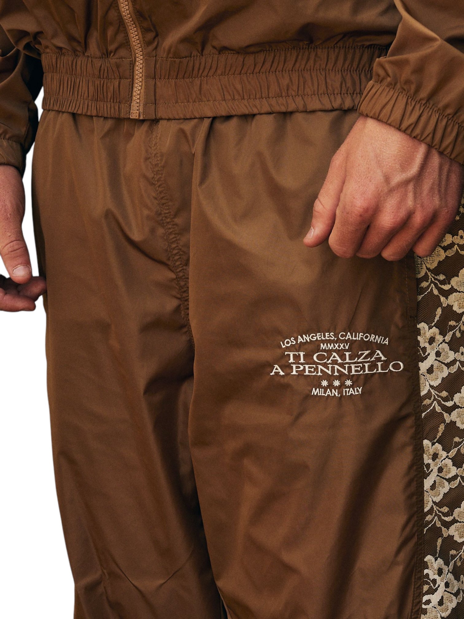 A close-up shows a person in Pleasures Francesca Track Pants Brown by PLEASURES, featuring polyamide lace details and white text reading "LOS ANGELES, CALIFORNIA MCMXCV TI CALZA A PENNELLO MILAN, ITALY," exemplifying Italian craftsmanship.