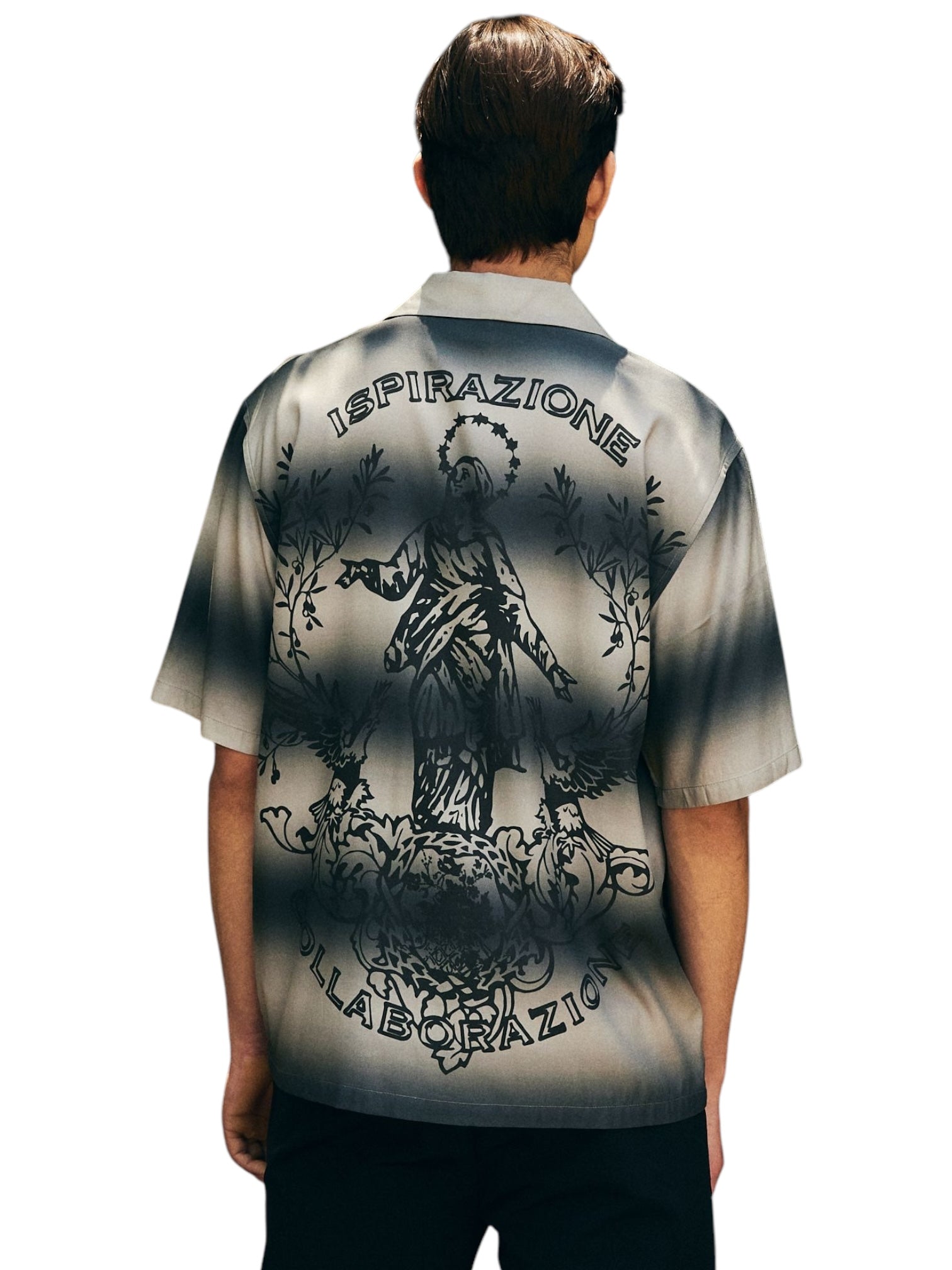 A man wears the Pleasures Giovanni BUTTON UP SILVER shirt by PLEASURES, featuring a graphic of a person among leaves with "ISPIRAZIONE" and "COLLABORAZIONE" on the back. It has a stylish camp collar, crafted from premium Viscose Challis, and is proudly made in Italy.