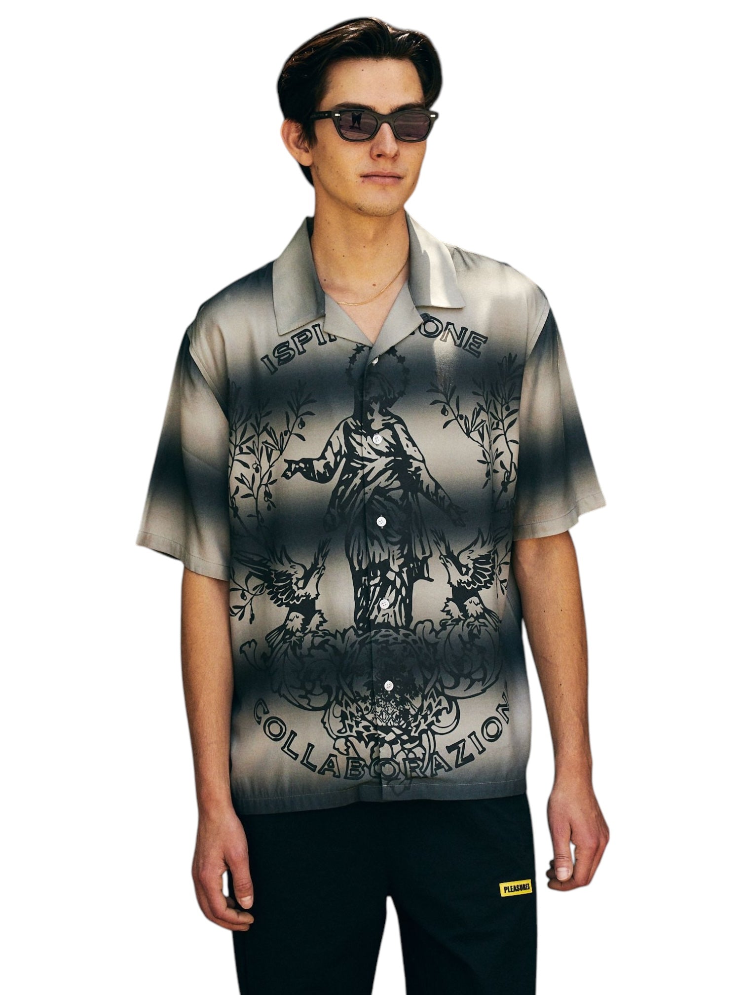 A man wearing the Pleasures Giovanni BUTTON UP SILVER by PLEASURES, with sunglasses and a patterned short-sleeve camp collar shirt made of luxurious Viscose Challis, featuring graphics and text. He pairs it with dark pants for a relaxed, stylish look, standing with his hands by his sides.