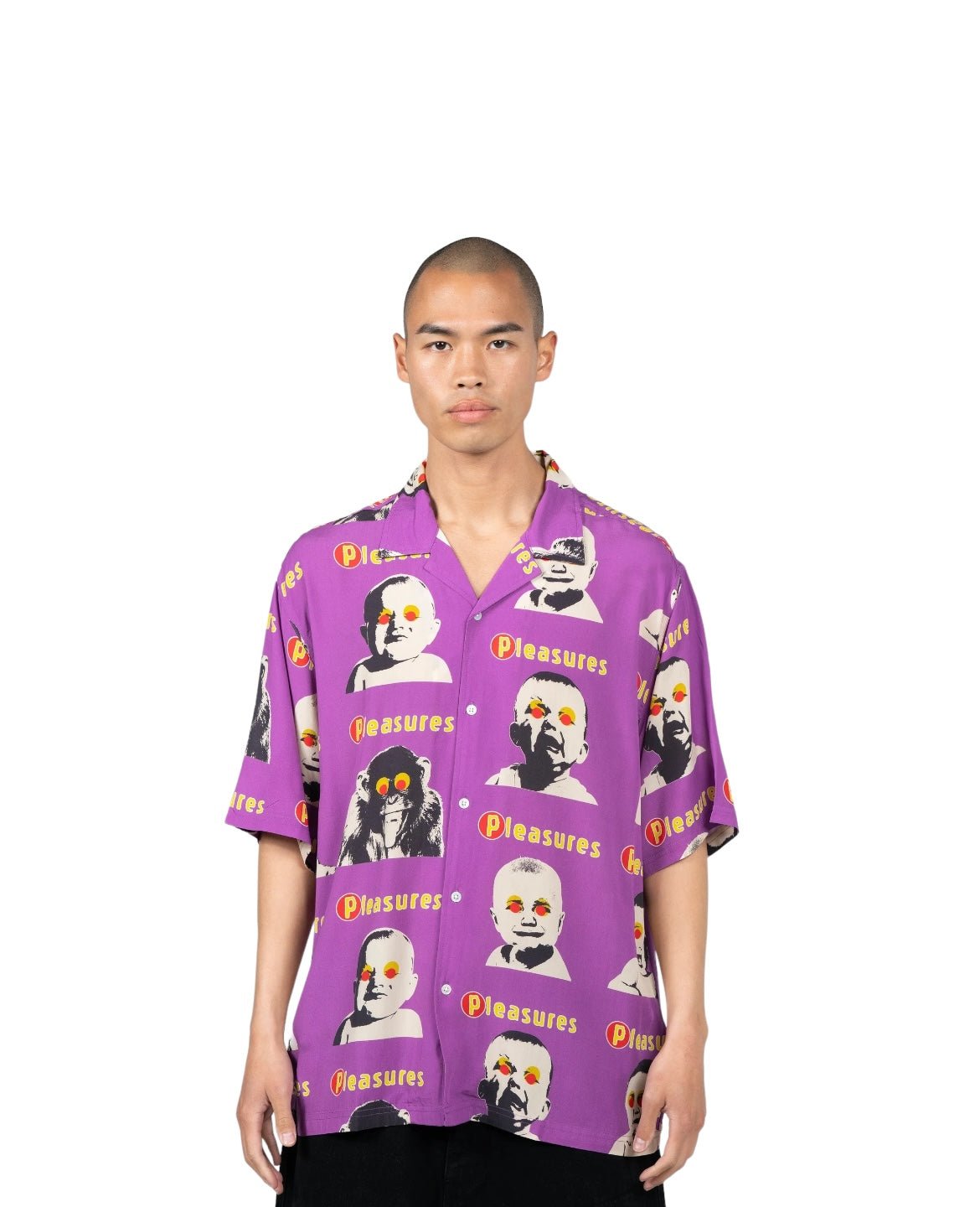 A person standing, wearing the PLEASURES HEAD BUTTON DOWN PURPLE with the word "PLEASURES" and graphic faces patterned across its sleek purple design.