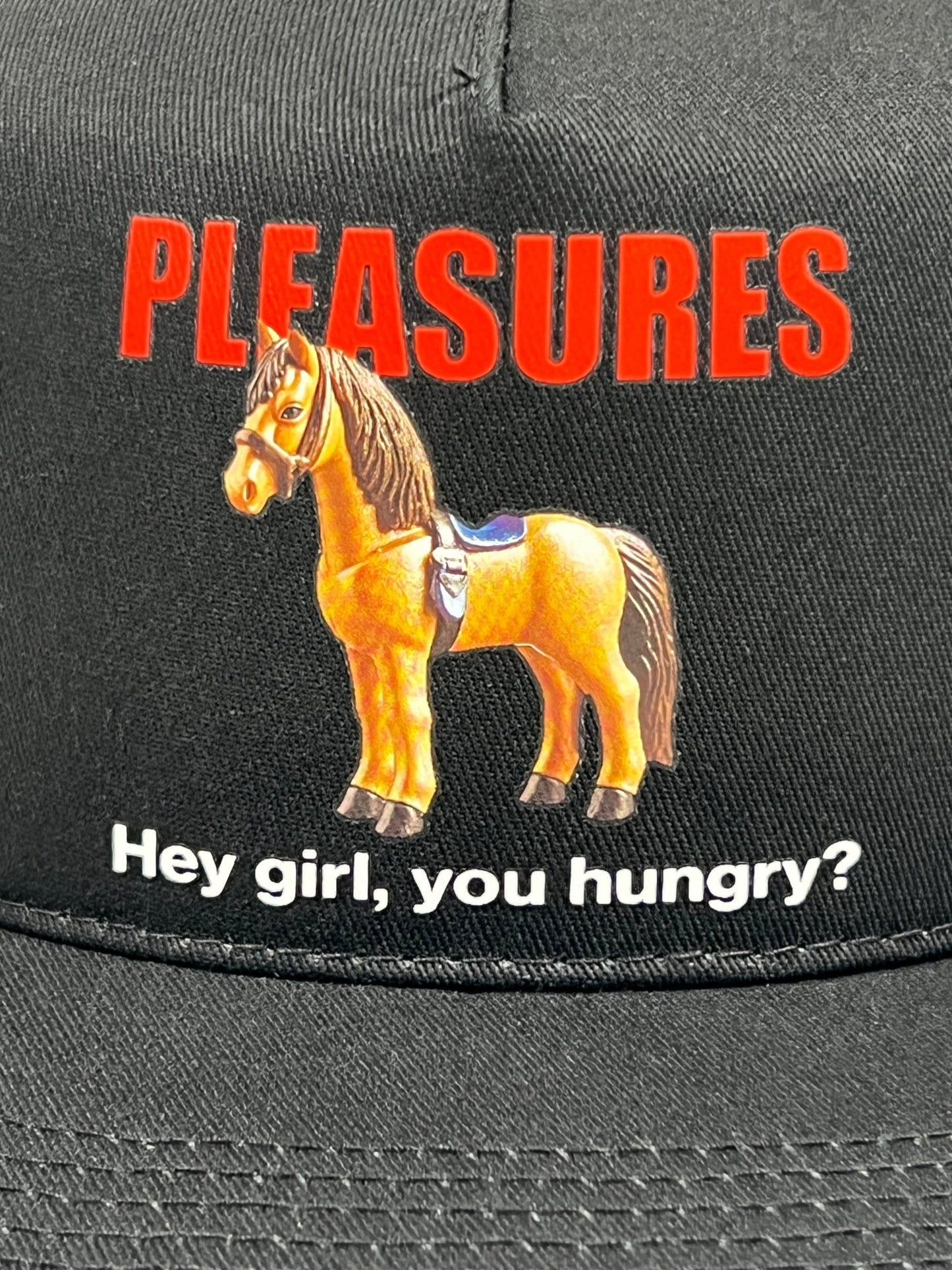 The PLEASURES HORSE TRUCKER BLACK hat by PLEASURES is crafted from black polyester and features red "PLEASURES" text above an image of a toy horse. Below the horse, white text reads, "Hey girl, you hungry?" on the front.