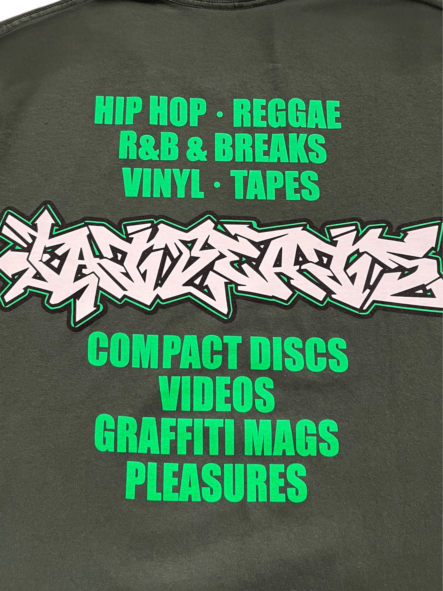 Green and white text on a dark background lists music genres, formats, and items—including hip hop, reggae, vinyl, tapes, CDs, videos, graffiti mags—featured on the Pleasures Melrose T-shirt Ivy by PLEASURES.