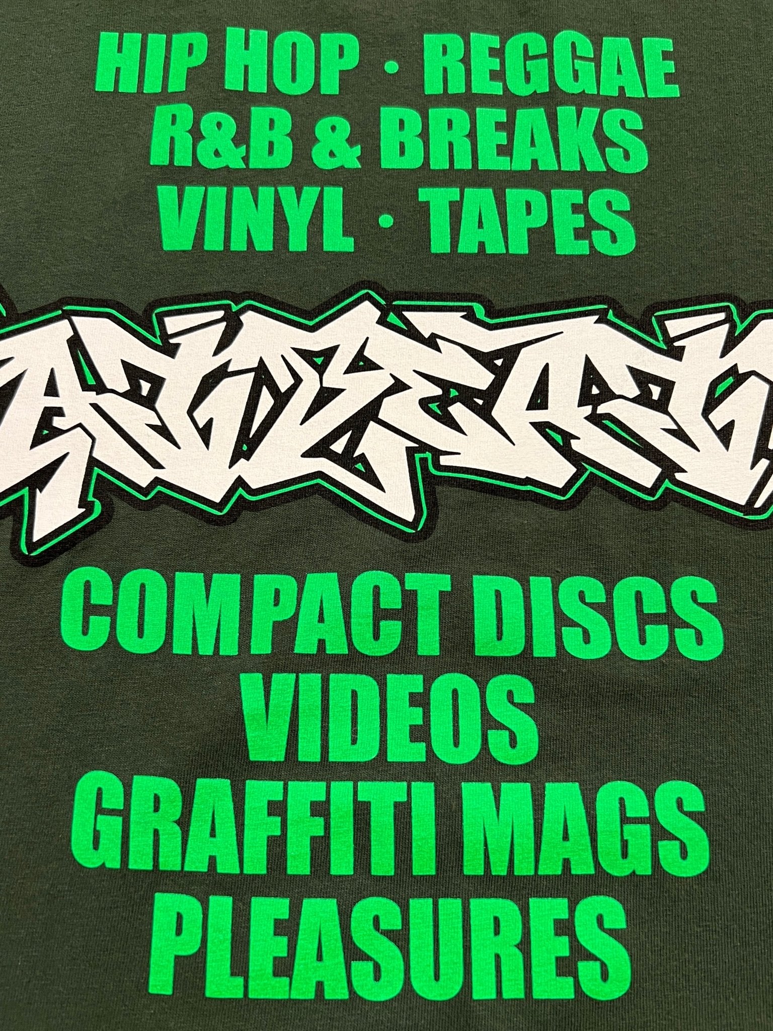 The PLEASURES Melrose T-shirt Ivy features green graffiti-style text on a dark background, listing music genres and media like hip hop, reggae, vinyl, tapes, and more. Made in the USA for true Fat Beats fans.