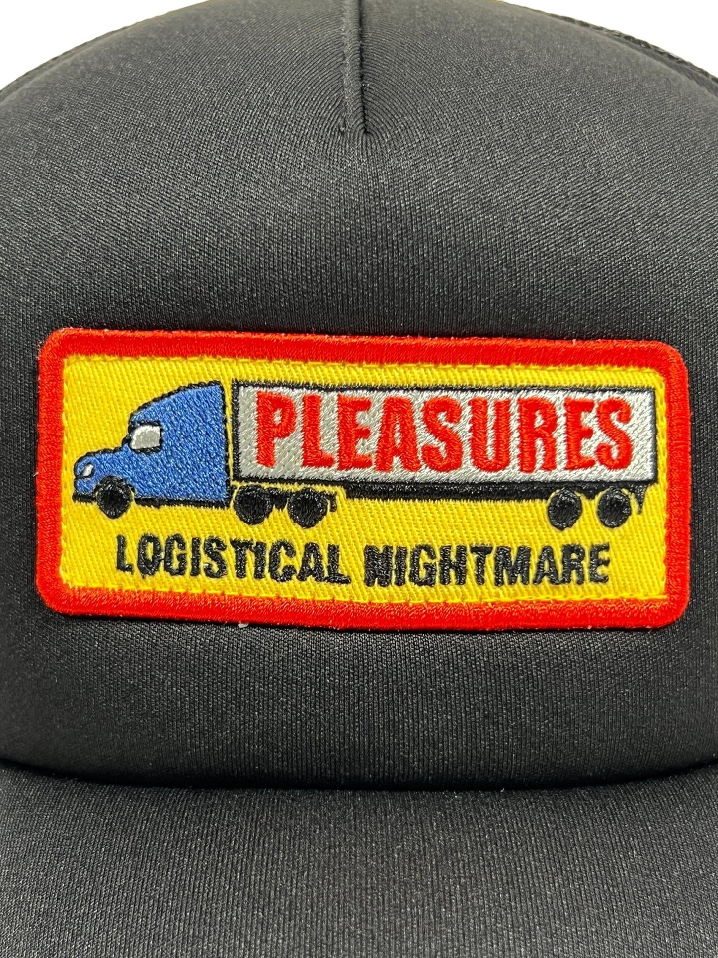 A close-up of a PLEASURES NIGHTMARE TRUCKER BLACK hat with a patch depicting a blue truck and trailer. The trailer has the text "PLEASURES," while below the image it reads "LOGISTICAL NIGHTMARE." The hat features durable polyester construction.