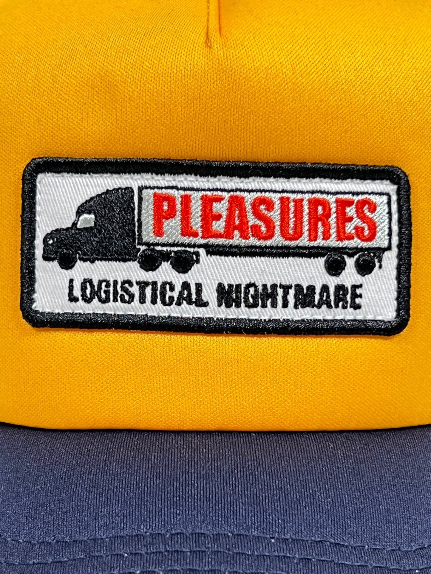 Close-up of the navy and yellow PLEASURES NIGHTMARE TRUCKER hat featuring an embroidered patch with a truck image, the word "PLEASURES" in red, and the phrase "LOGISTICAL NIGHTMARE" in black. Crafted from durable nylon for lasting wear.