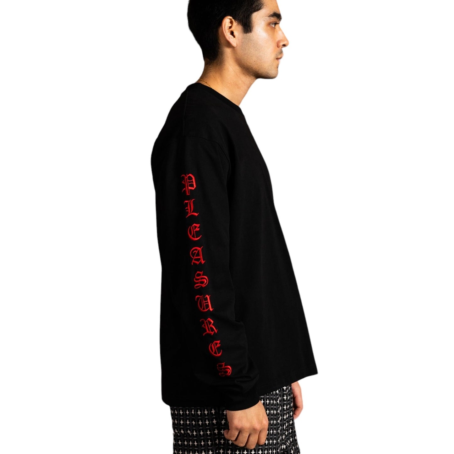 A person is wearing the Pleasures Oe Heavyweight Long Sleeve Black shirt, featuring "PLEASURES" in red satin stitch embroidery along the sleeve, paired with patterned pants.