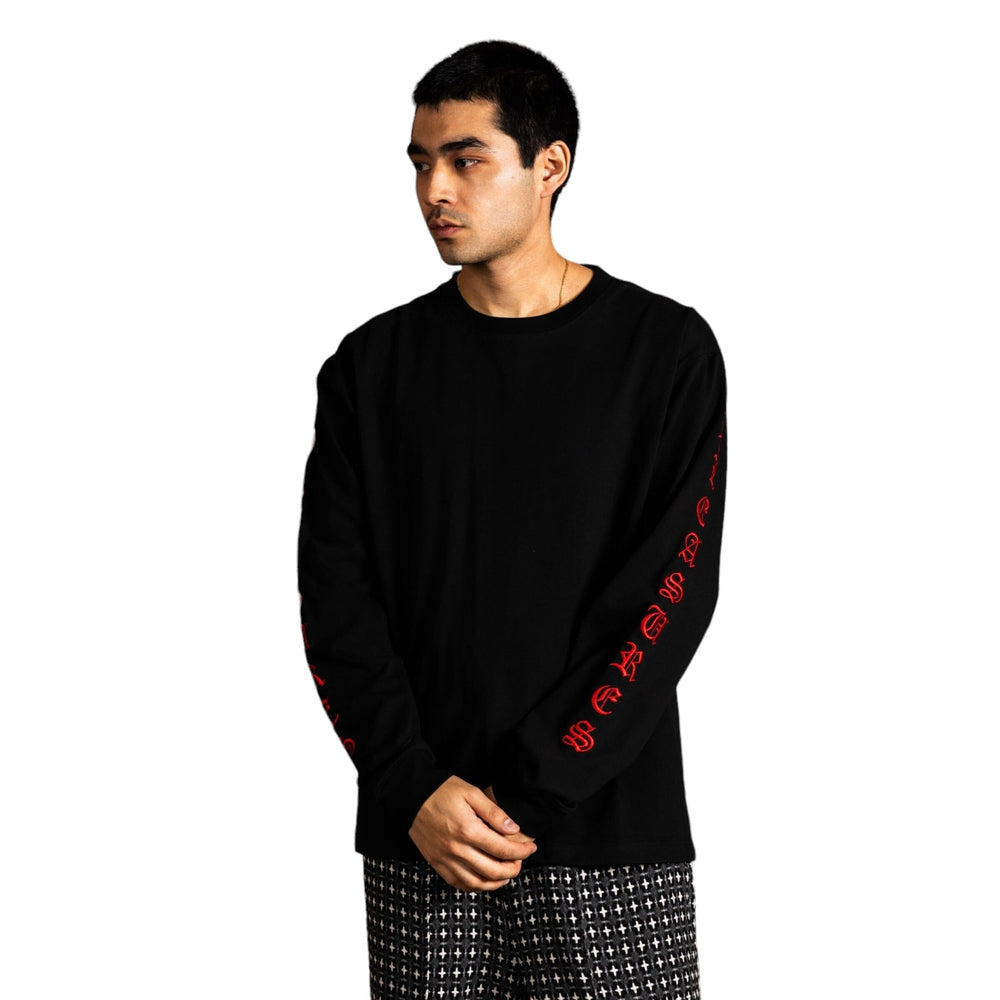 A man in a PLEASURES Oe Heavyweight Long Sleeve Black shirt, featuring bold red text on the sleeves, paired with patterned pants, stands confidently against a white background.