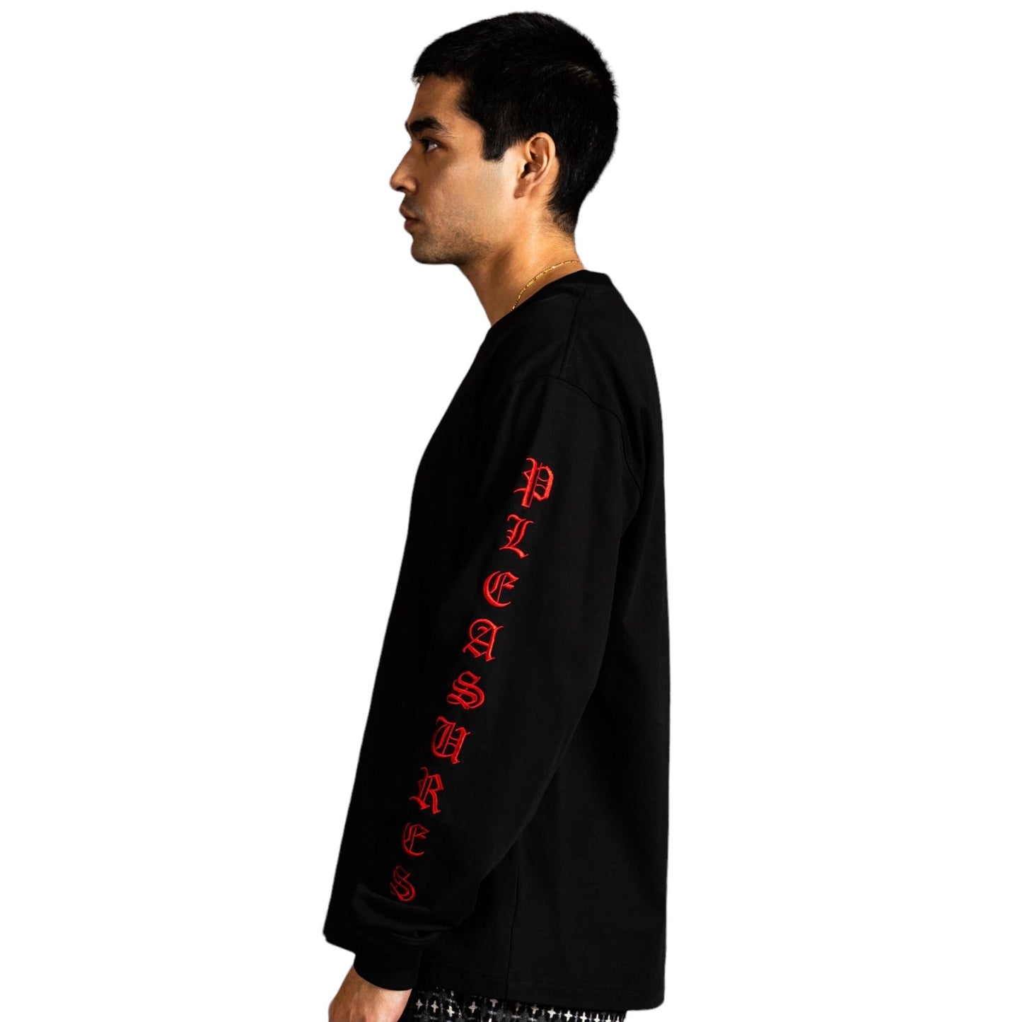 A side view showcases a person wearing the Pleasures Oe Heavyweight Long Sleeve Black shirt, featuring bold red satin stitch embroidery of "PLEASURES" on the sleeve.