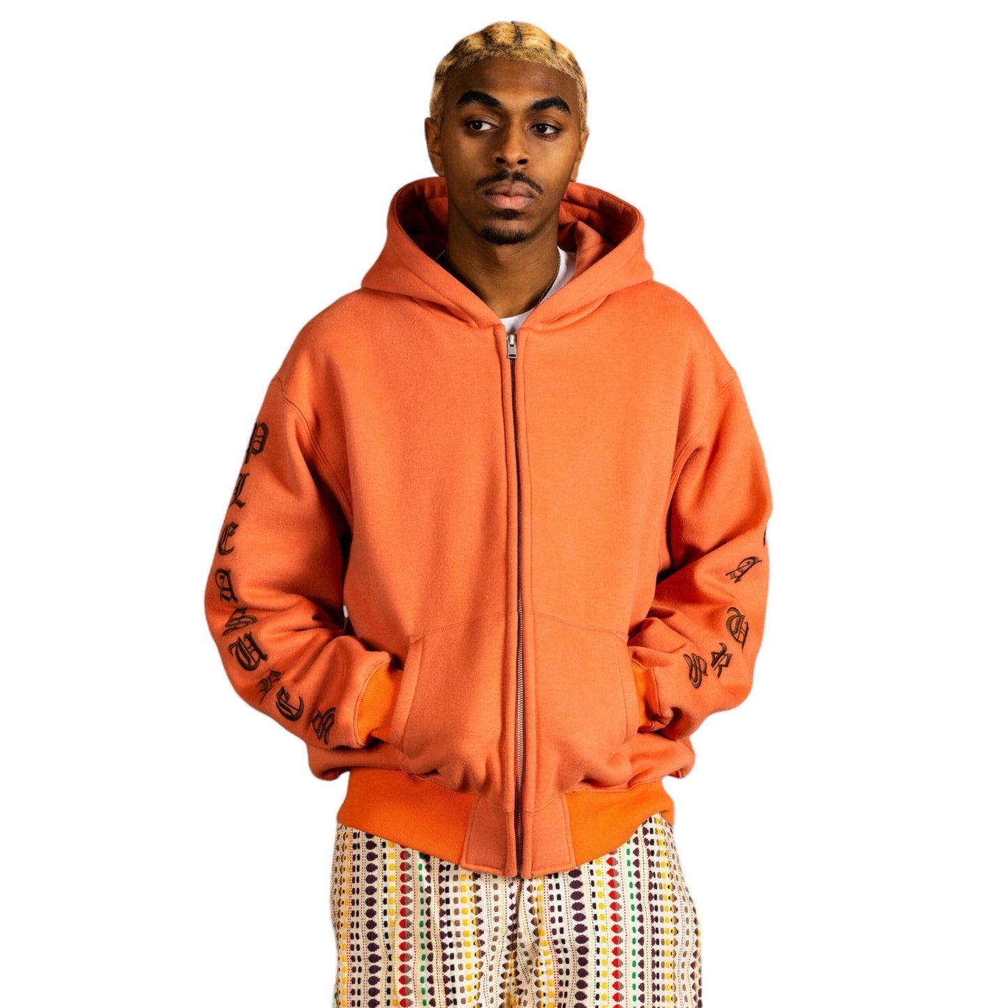 A person wearing the PLEASURES OE ZIP UP HOODIE RUST, an orange hooded jacket with black text on the sleeves, paired with patterned pants, stands against a white background. This relaxed fit hoodie from PLEASURES is made from heavyweight cotton, offering both style and comfort, perfect for any casual outing.