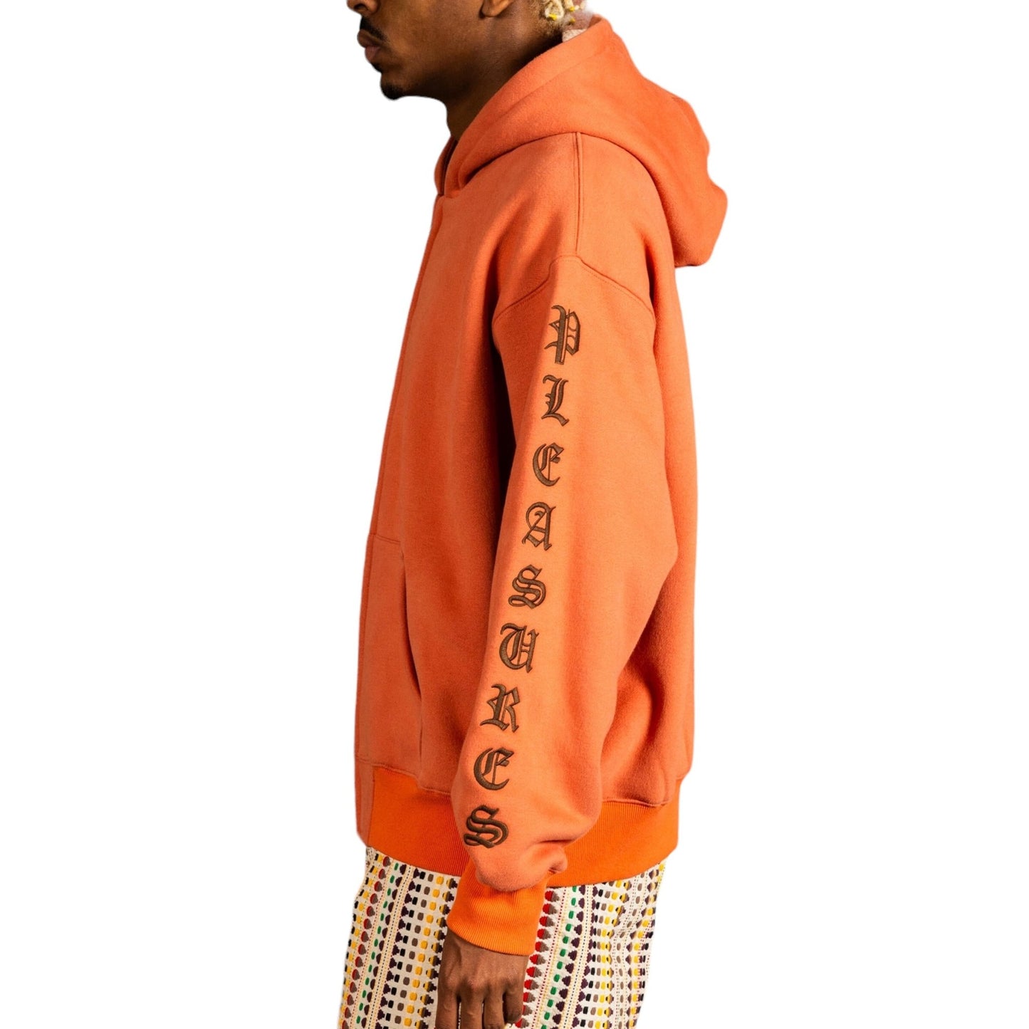 A person wearing the PLEASURES OE ZIP UP HOODIE in rust, with "PLEASURES" written down the sleeve and multicolored patterned pants, facing sideways.