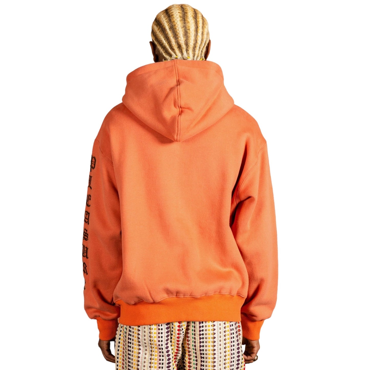 A person with braided hair is seen from the back, wearing a relaxed fit PLEASURES OE ZIP UP HOODIE RUST made of heavyweight cotton and patterned pants. The hoodie, featuring text on the right sleeve, appears cozy with its brushed fleece lining.