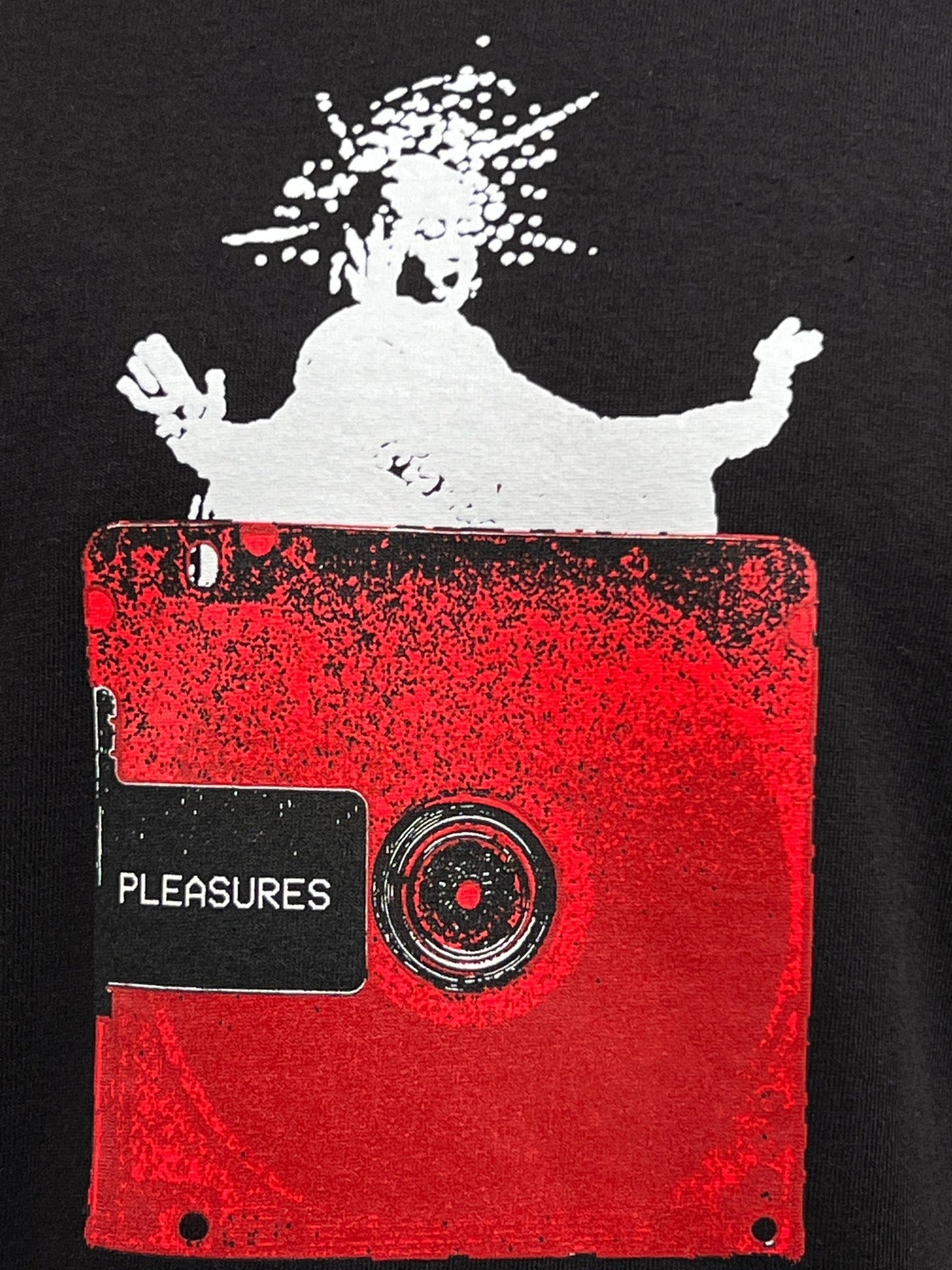 A white silhouette figure on top of a red square object with a black strip labeled "PLEASURES." The background is black. This design graces the front of the PLEASURES OPERA T-SHIRT BLACK from PLEASURES, ensuring comfort and breathability with its pure cotton, crewneck style.