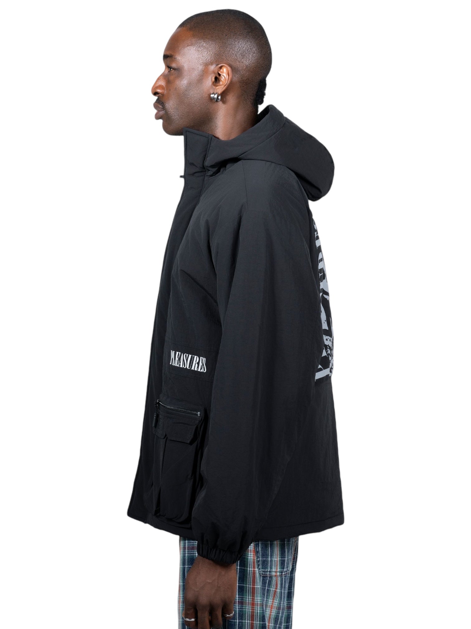 Man in profile wearing a Pleasures Run Hooded Jacket in Black from PLEASURES, featuring graphic text on the back and sleeve, crafted from 100% nylon, paired with plaid pants.