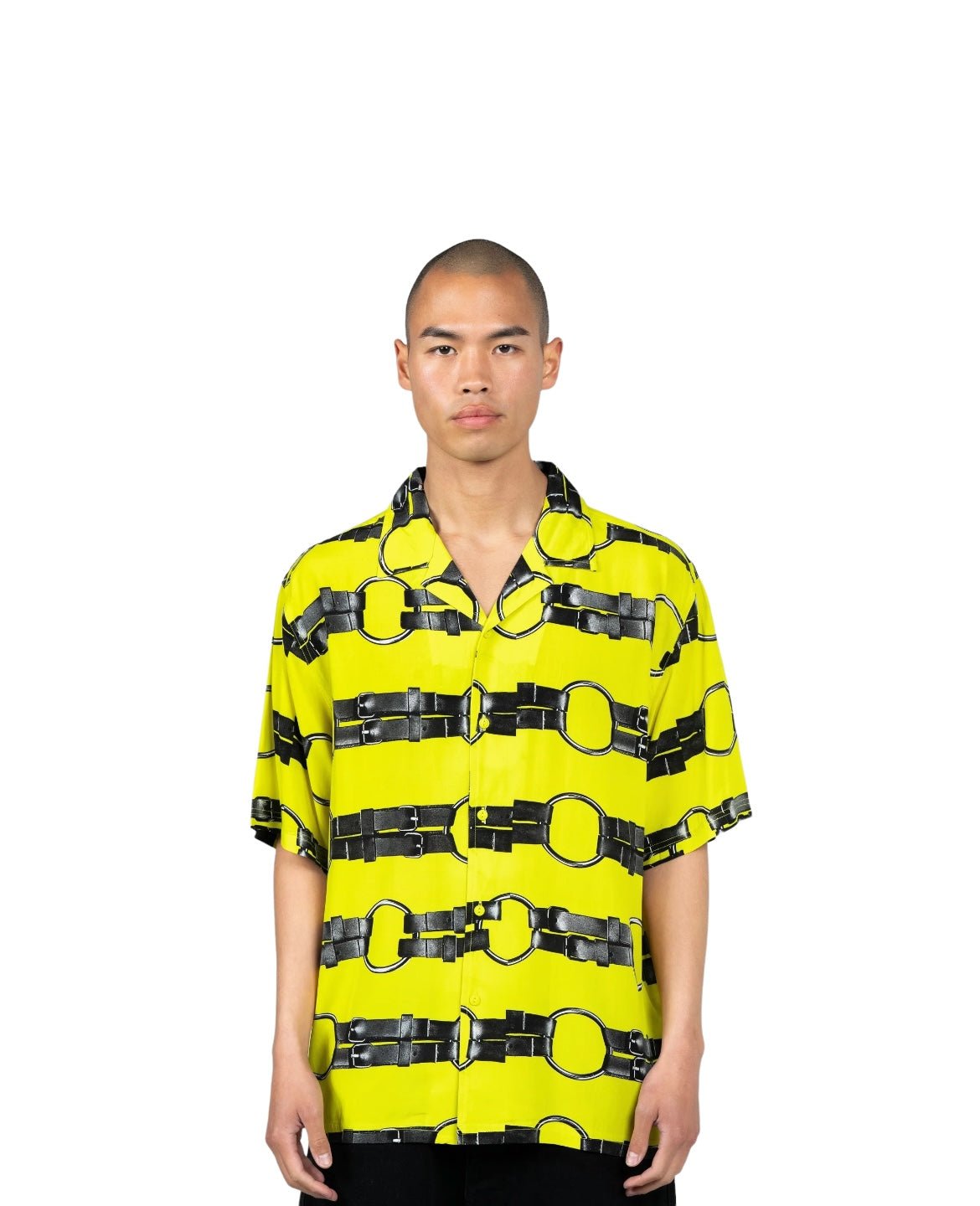 A person is wearing the PLEASURES STRAPS BUTTON DOWN YELL, a bright yellow, camp collar short sleeve button-up shirt with a black belt pattern, standing facing the camera against a white background. The shirt is by PLEASURES and features vintage 2-hole buttons.