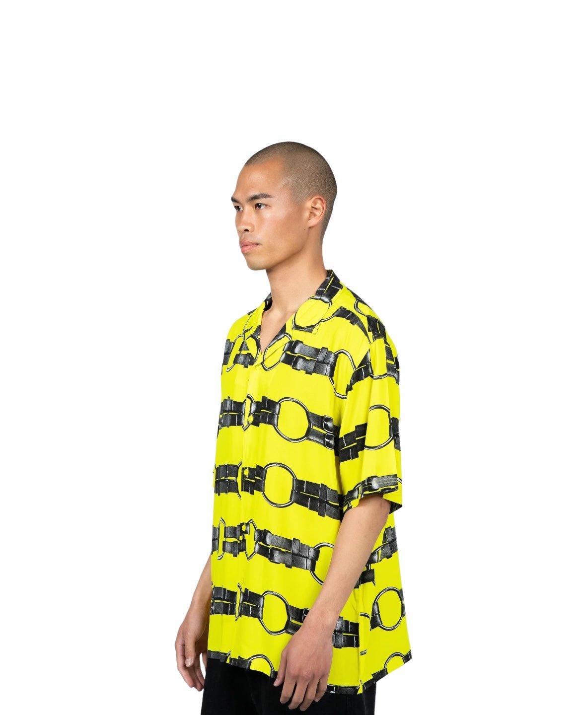 A person stands facing left, wearing the PLEASURES STRAPS BUTTON DOWN YELL, a vibrant yellow short sleeve shirt with a camp collar and a striking black chain pattern. The background is plain white.