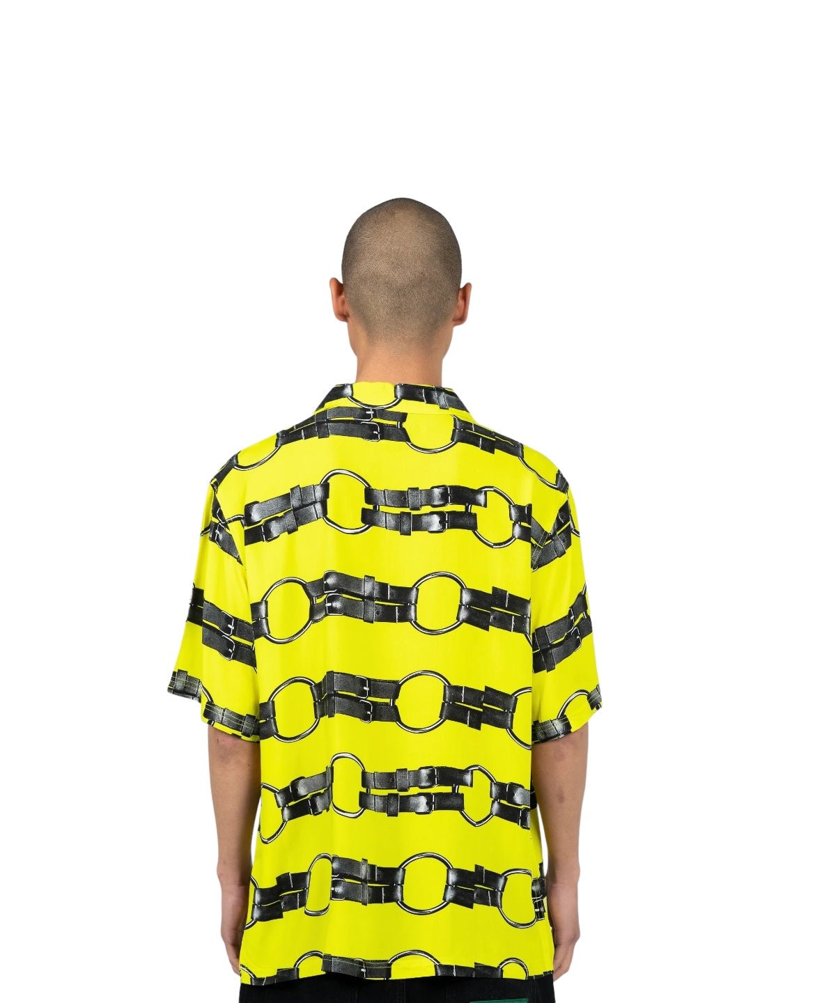 A person with a shaved head is seen from behind wearing the PLEASURES STRAPS BUTTON DOWN YELL, a bright yellow shirt with a black chain pattern. The camp collar short sleeve button-up design, adorned with vintage 2-hole buttons, stands out crisply against the plain white background.