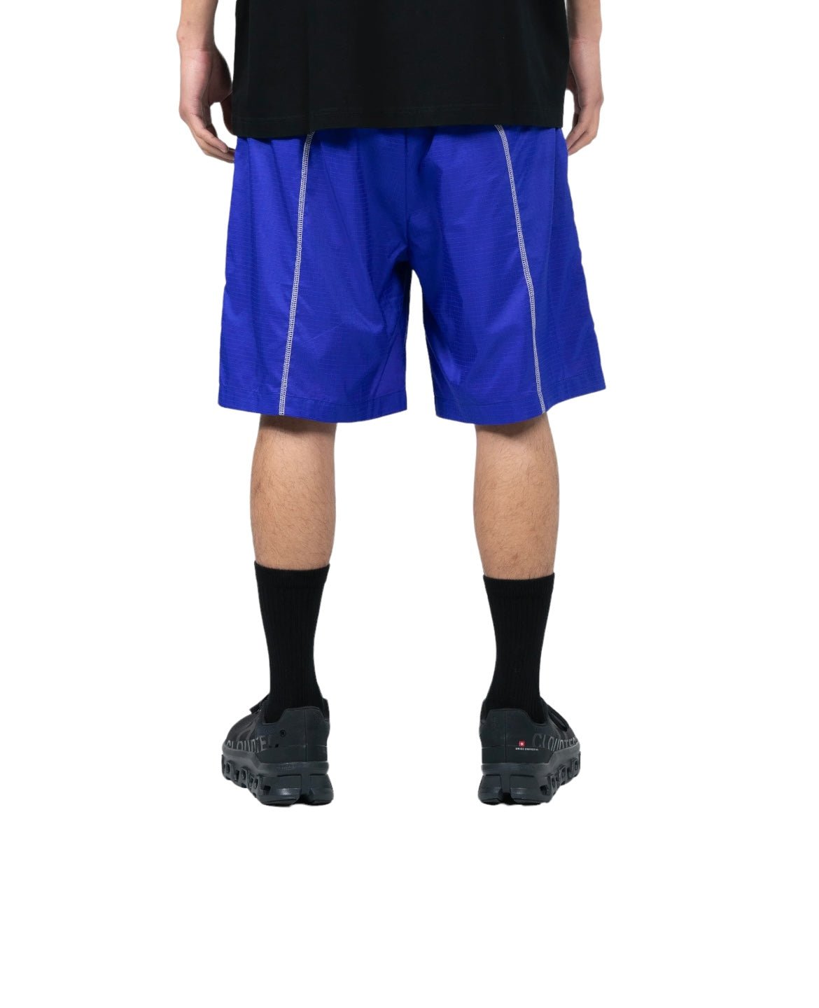 Person standing and facing away from the camera, wearing blue PLEASURES TEMPO ACTIVE SHORTS BLUE, black socks, black shoes, and a black shirt. The lightweight durable shorts are perfect for an active lifestyle.